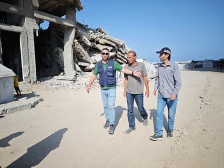 Three men, one with a blue WFP vest, on a dusty road in front of a collapsed raw concrete building.