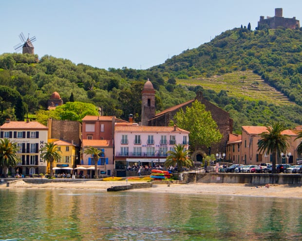 A small town of pastel buildings alongside a sandy shore and backed with green hills with a fort and a windmill on them