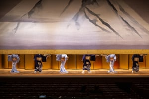 Six geishas on stage, their heads hidden behind a part-raised curtain