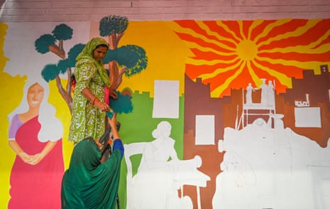Two women work on a colourful unfinished mural depicting a bright sun, people and housing