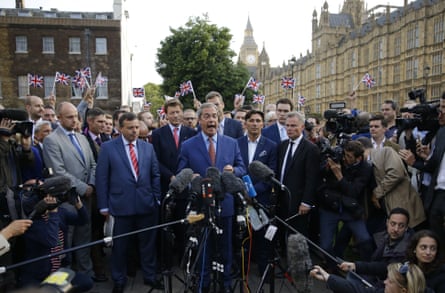 Nigel Farage surrounded by a crowd of people waving union flags and holding microphones.