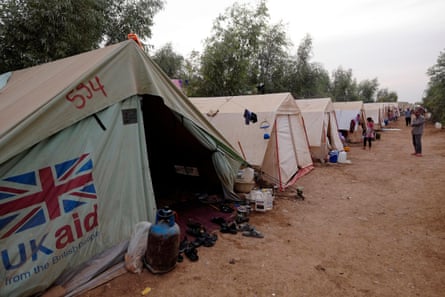 a union jack flag and the words UK aid from the British people are seen on the side of the tent closest to the camera. There are shoes, water containers and cooking gas cylinders at the tent's opening.