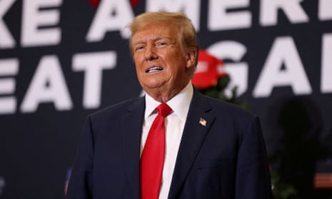 Older white man, orangey skin, poofy hair, in dark suit, red tie, white shirt on stage with partial letters of 'Make American Great Again' visible.