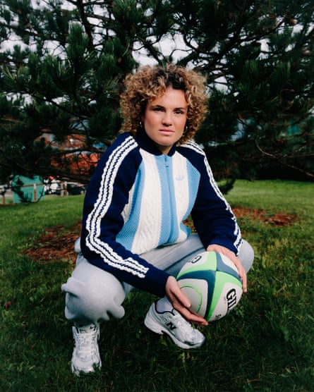 Ellie Kildunne crouching down holding a rugby ball wearing a knitted tracksuit top with a pale blue and white thick-striped front and black arms with white Adidas stripes down the length, grey tracksuit bottoms and white socks and trainers