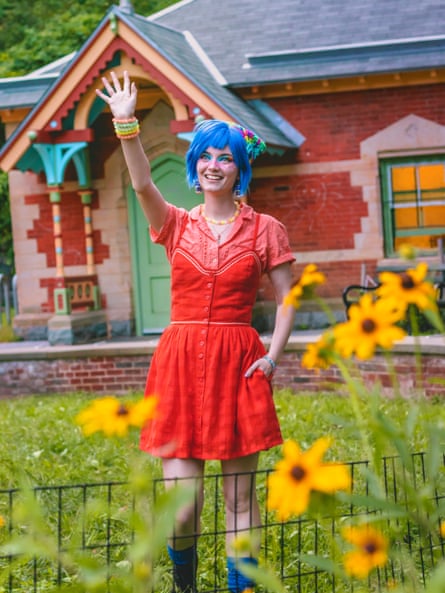 Lizard Leigh with blue hair and a red dress, standing in front of a colourful house