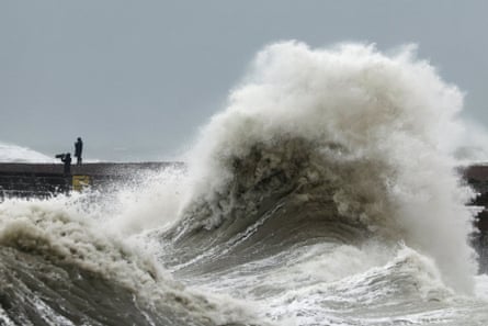 A huge wave crashes over Newhaven Harbour wall as two people watch from a distance