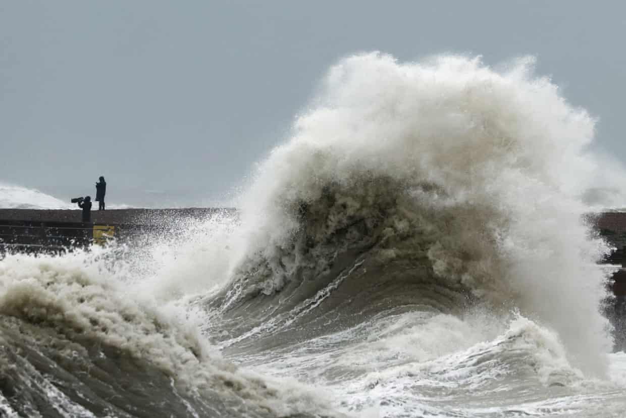A huge wave crashes over Newhaven Harbour wall as two people watch from a distance