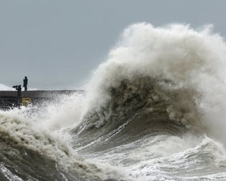 A huge wave crashes over Newhaven Harbour wall as two people watch from a distance