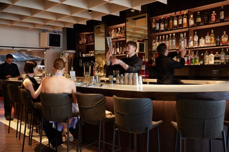 A bartender pours a cocktail by upturning a bottle into a glass behind a bar for two customers sit on stools at the bar