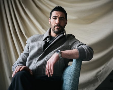 The actor Tahar Rahim looking serious, wearing a light grey jackeet, sitting relaxed on a blue chair.
