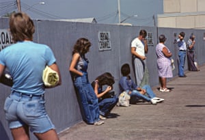 a Willy Spiller photo titled "Summer in Queens, 1983": people stand next to a wall waiting in line for something on a sunny day