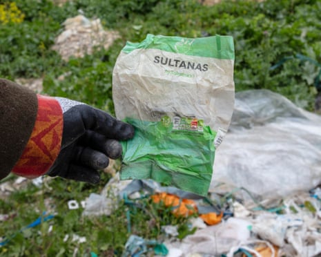 A gloved hand holding a crumpled sultana packet from Sainsbury's amid dumped plastic waste.