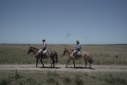Two men ride horses along a track on a plain, in the background are several wind turbines.
