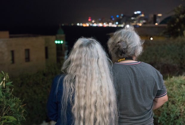 Two women with grey hair photographed from behind outside at night with the lights of a city in the distance