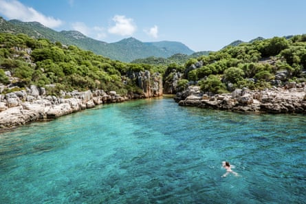 A woman swimming in very clear turquoise water towards a small cove on a rocky shoreline