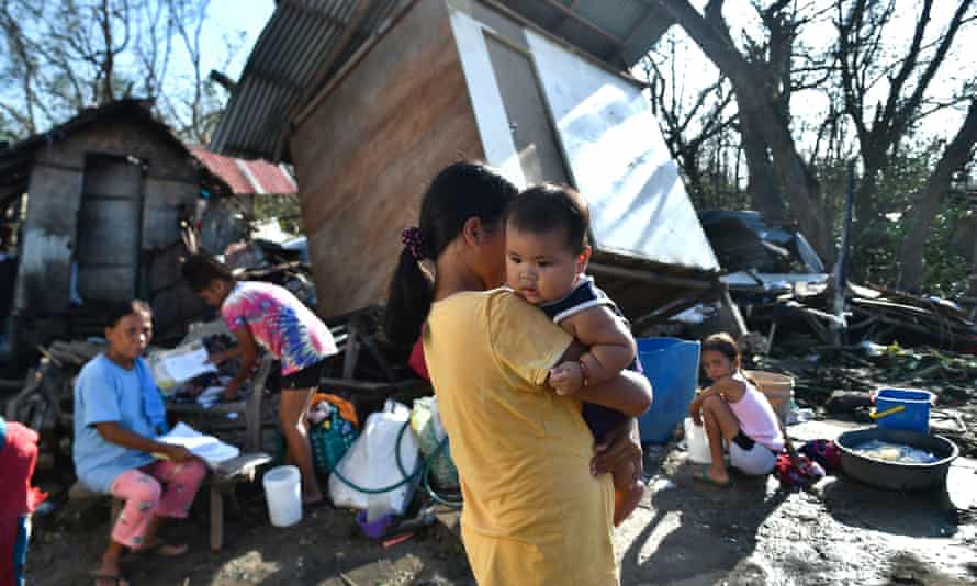 A woman holds a baby while an older woman and a child sit in the ruins of their house