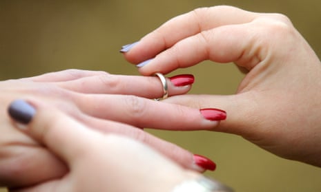One white hand, with blue painted fingernails, places a gold band on the ring finger of another white hand, with red fingernails.