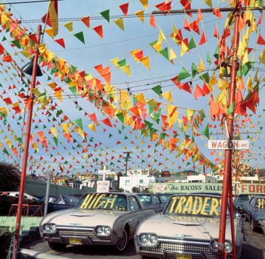 The forecourt of a used car trader shows two cars in a space where red poles hold up coloured bunting above them. On the windscreen of one, large yellow letters say ‘HIGH’ and on the windscreen of the other ‘TRADER’