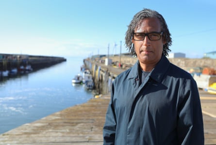 A man in a blue raincoat standing by a pier with the sea behind him