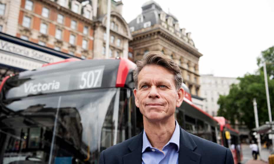 Head and shoulders shot of David Brown in a suit and open-necked shirt standing in front of a red number 507 single-decker bus, with the facade of Victoria station in the background