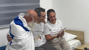 Dani Miran, draped in an Israeli flag, has his arm on the shoulder of his son Omri as they look at a tablet being held by Omri’s wife Lishay as all three sit on a single bed