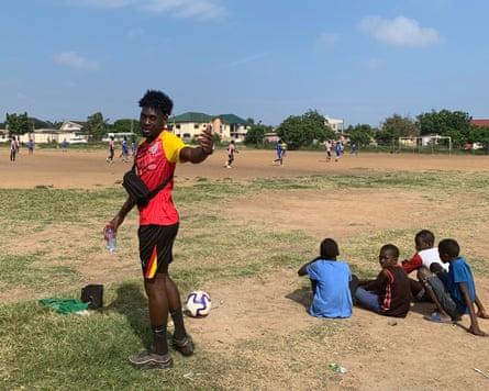 Gabriel Oyewole standing beside a football pitch with four boys sitting on the grass next to him