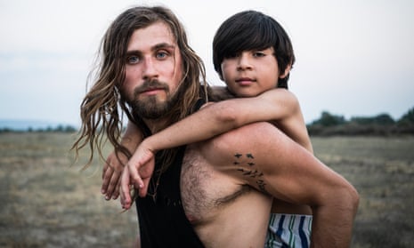 Alex, bare-chested, blue-eyed, long haired and with a beard, carrying a refugee boy on his back in open landscape