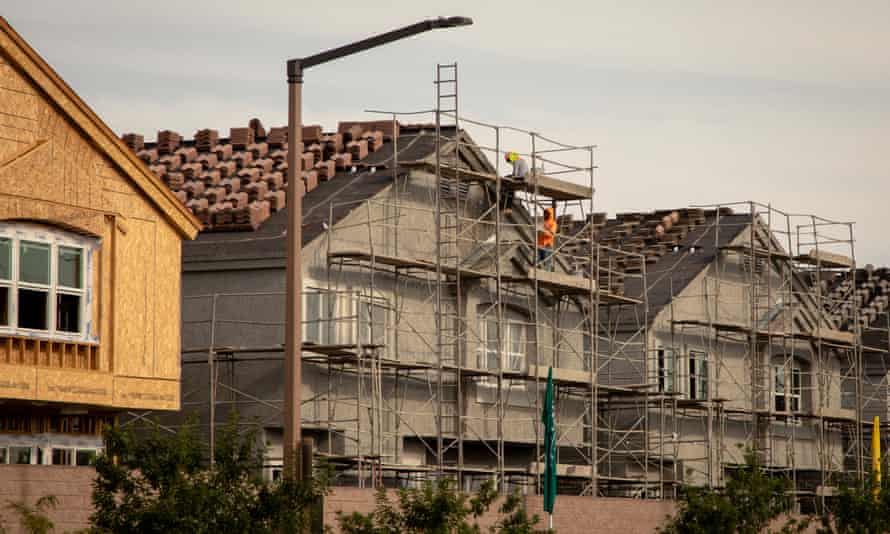 Construction workers climb up scaffolding as they work on the exterior of a newly built multi-story house. Houses in varying stages of completion are seen on either side. 