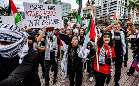 A group of people wearing red, white and black wave Palestinian flags, one holding a sign that says ‘Israel has killed +500 children in the past week.’