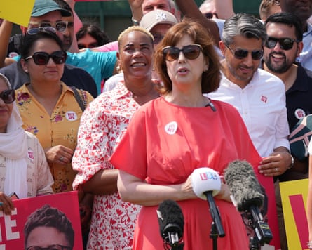 Lucy Powell speaks at an event: she is wearing a bright red dress with elbow-length loose sleeves and dark sunglasses. She stands in front of a crowd in the sunshine.