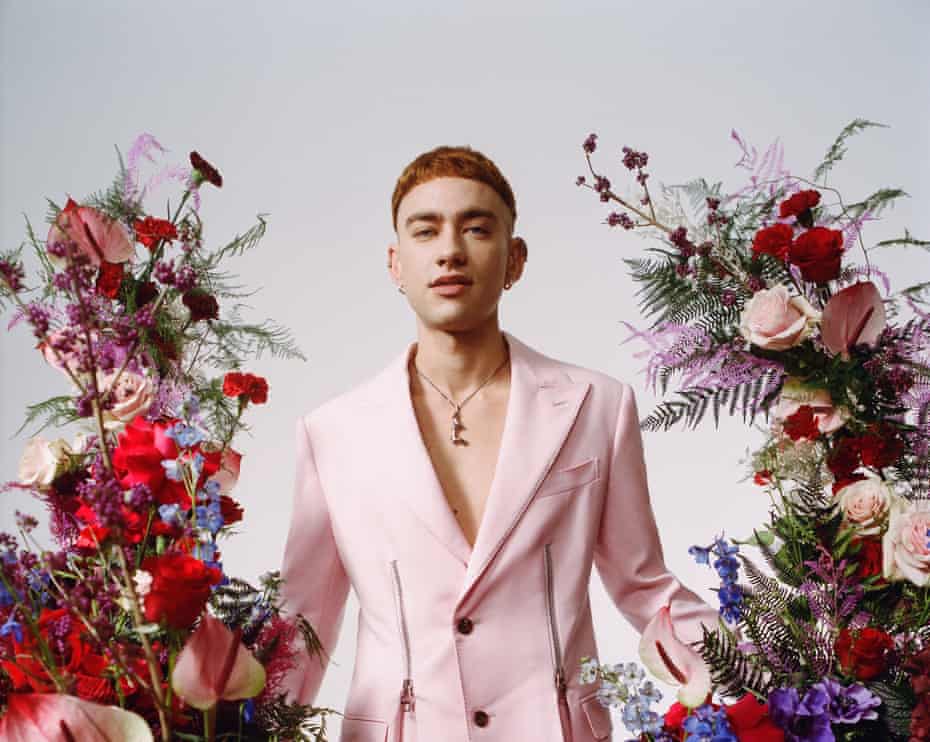 Olly Alexander wearing a pink Alexander McQueen suit and surrounded by pink, red and purple flowers