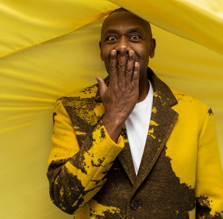 Lenny Henry in a yellow and brown jacket and white T-shirt, against a billowing background of yellow fabric