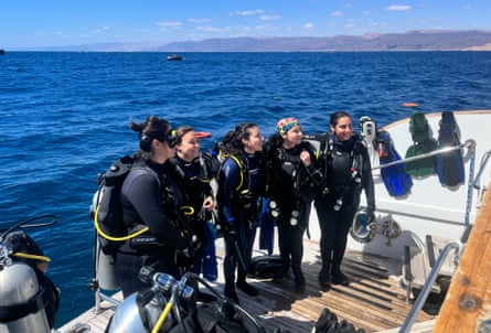 Five female scuba divers in wetsuits and with air tanks on a boat’s platform