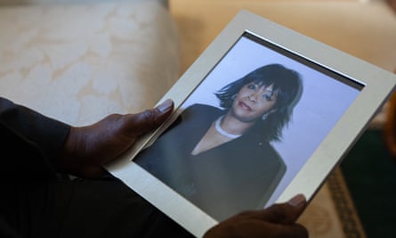 Two black hands hold a silver-framed photo of a black woman with black bangs and chin-length hair, wearing a black jacket and a white necklace.