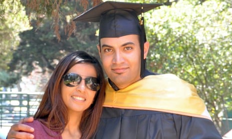 A woman with long brown hair and black aviator sunglasses stands outdoors beside a taller man wearing a black graduation cap and black robe with a gold-colored mantle, his arm around her shoulders and her leaning into him. Both appear Middle Eastern and both are smiling.