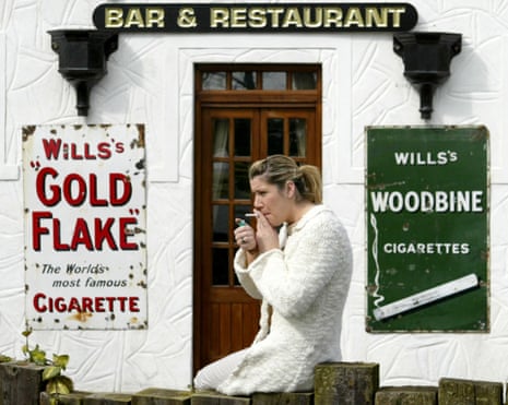 A woman wearing a white coat smokes a cigarette outside a bar