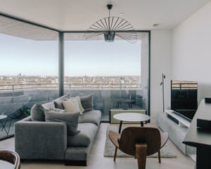 A modernist grey sofa, a three-legged wooden occasional chair and small round coffee table in the living room of an apartment with floor to ceiling windows