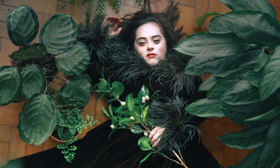 A woman with Down's syndrome in a black velvet dress with a furry collar, lying on her back with green leaves around and on her