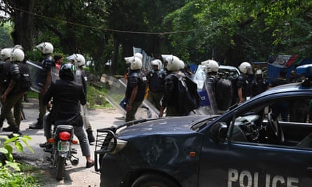 A dozen police officers wearing helmets and carrying shields gather on a road in front of a police vehicle.