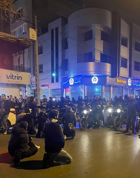 Young men crouch and kneel in a street facing a line of police on motorbike; other people are standing by in front of shops.