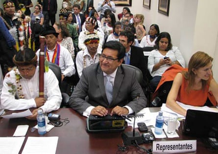 A Latino man in a suit sits at desk next to an Indigenous man wearing feathers in his hair, with other similarly dressed people behind them