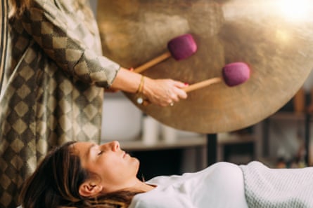 A person playing a gong in sound bath healing therapy, with a woman lying down nearby with eyes closed
