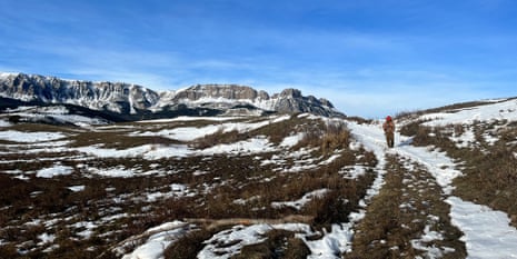 A person in an orange hunting cap and vest walks along a snow-dusted trail through open hills, with a mountain range in the background under a clear blue sky.