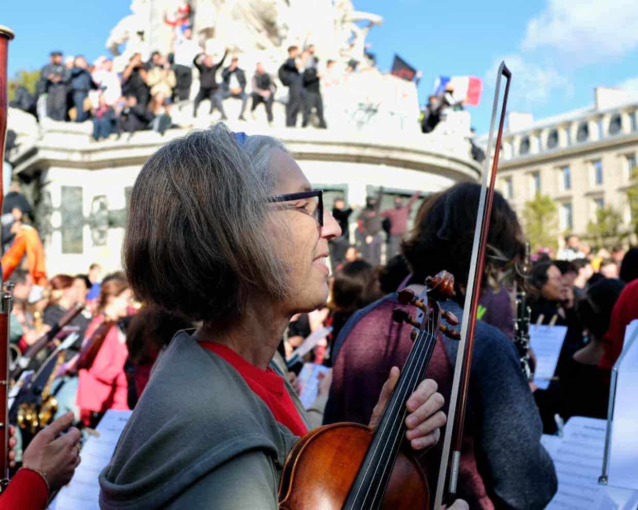 A smiling violinist smiles among her fellow orchestral players outdoors at Place de la République.