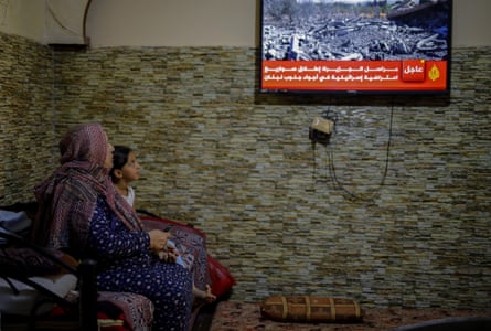 An older Arab woman in a headscarf sits next to a girl watching news about the war in Gaza on a TV.
