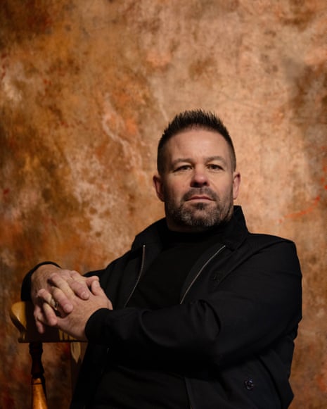 Gary Williamson sits in front of a marbled backdrop, facing the camera, with his clasped hands to the side, leaning on the back of a wooden chair
