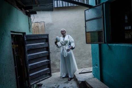 A black woman wearing a white dress and head scarf stands in a dark corridor