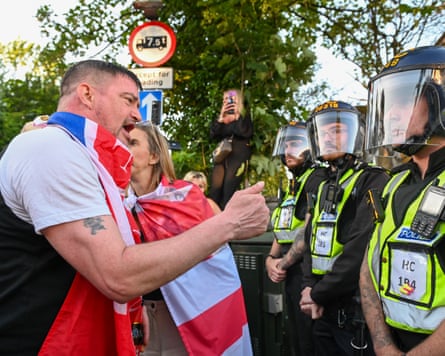 Man with a union flag around his shoulders gestures at a line of police officers with protective helmets on