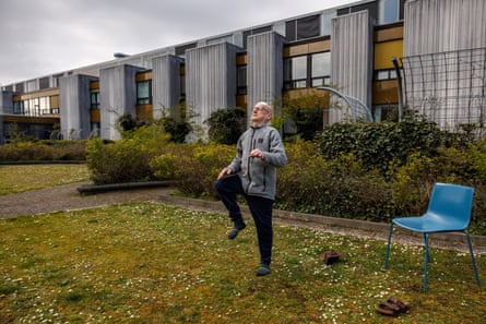 A man in a fleece jacket, leggings and socks stands on a lawn outside a two-storey hospital building exercising by lifting his right leg, hand on his knee