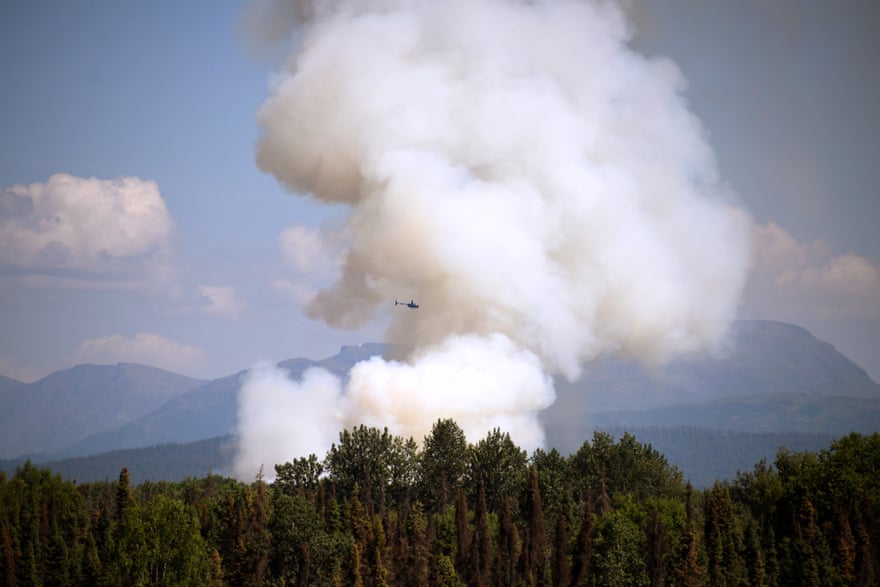 A helicopter is a small point in the sky next to the immense column of smoke that rises from a forest fire.